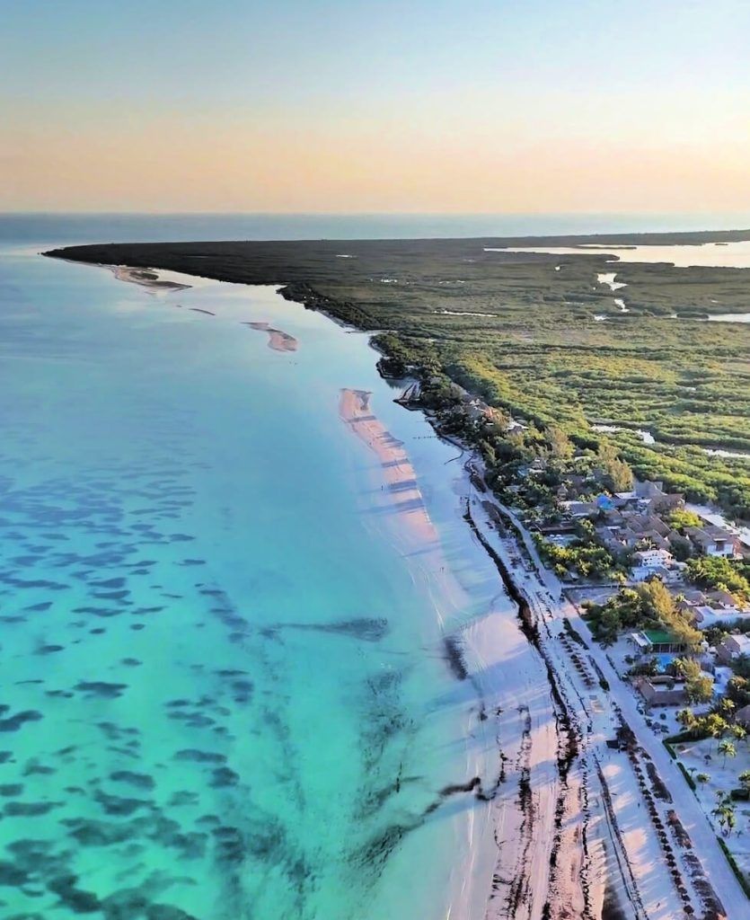 Aerial view of Holbox Island showing a clear water shoreline next to a sandy beach, with a dense area of vegetation further inland. The lighting suggests the photo was taken around sunset.