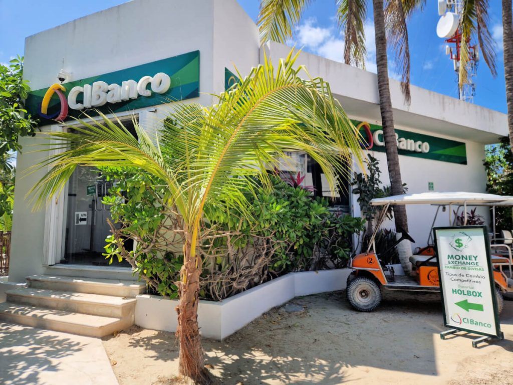 CiBanco bank branch in Holbox with an ATM entrance, flanked by tropical plants and a parked golf cart advertising money exchange services.