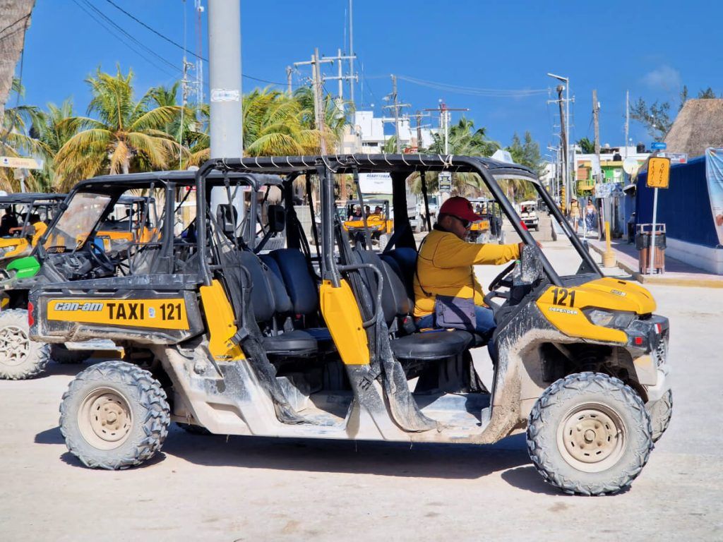 Golf cart taxi number 121 with a driver wearing a yellow shirt, parked on a sandy street in Holbox near the ferry port.