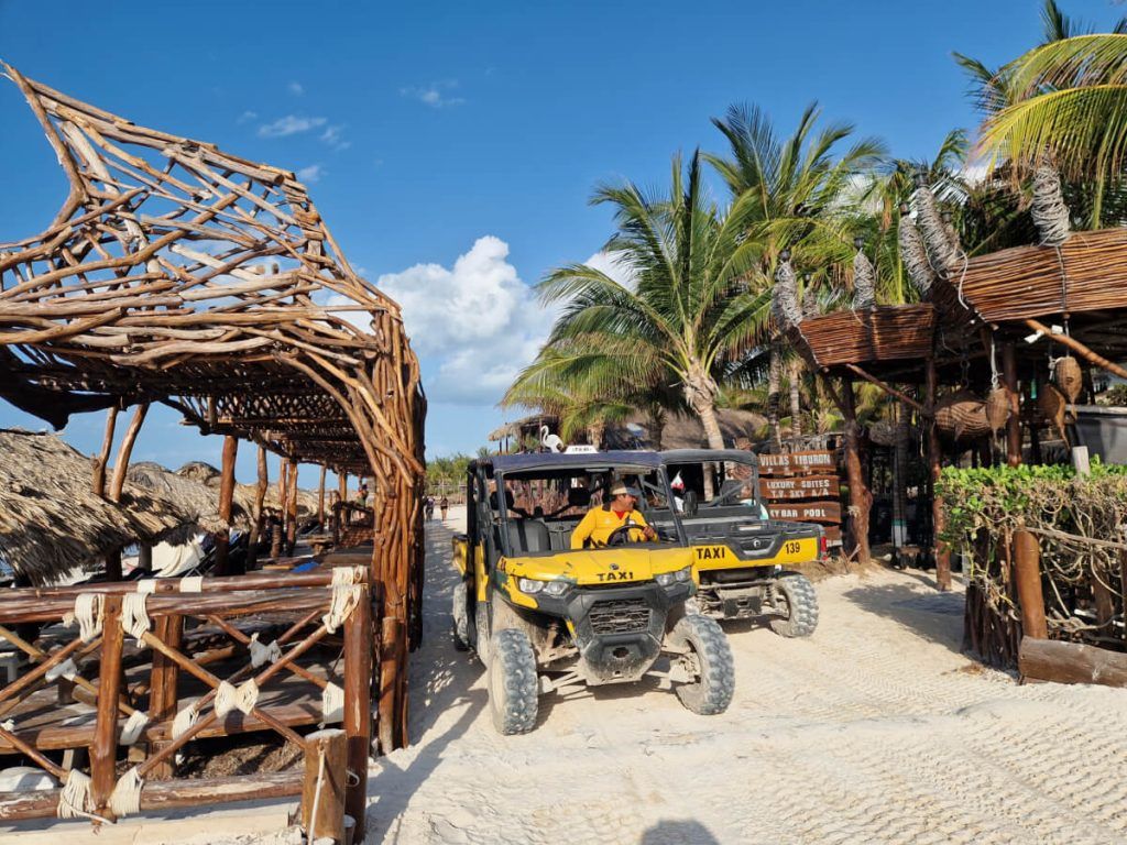 Golf cart taxis driving on a sandy beach road flanked by unique wooden structures and lush palm trees in Holbox.