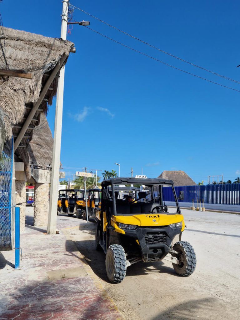 Front view of a parked yellow and black golf cart taxi with a 'TAXI' sign on its front, in Holbox with thatched structures in the background.