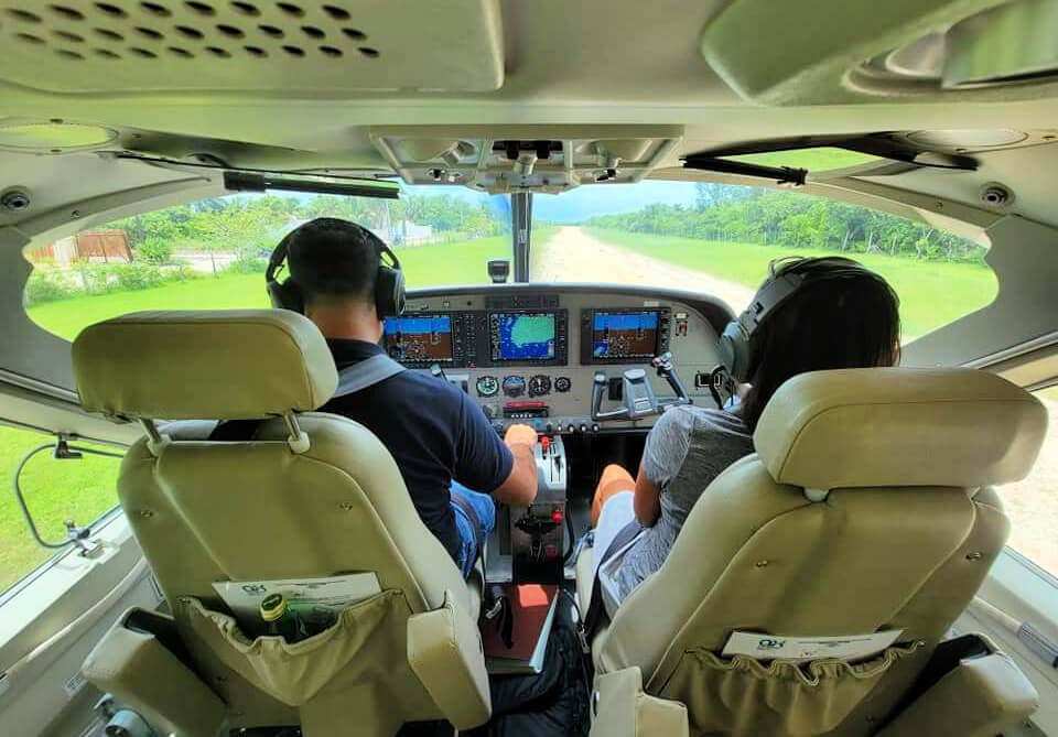 Interior view of a small aircraft cockpit with pilots wearing headsets during landing, with the runway ahead visible through the windshield, capturing the final moments before touchdown at Holbox Airport.