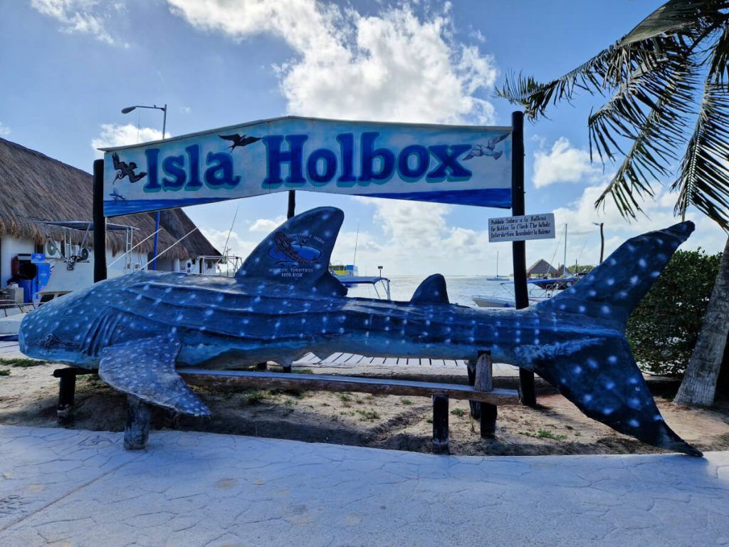 Sculpture of a whale shark in front of the 'Isla Holbox' sign at the ferry port, with palm trees and boats in the background.