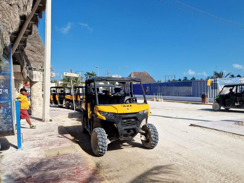 A line of yellow and black ATVs serving as taxis on a sandy street in Holbox, with a pedestrian nearby; tipping is appreciated for good service.