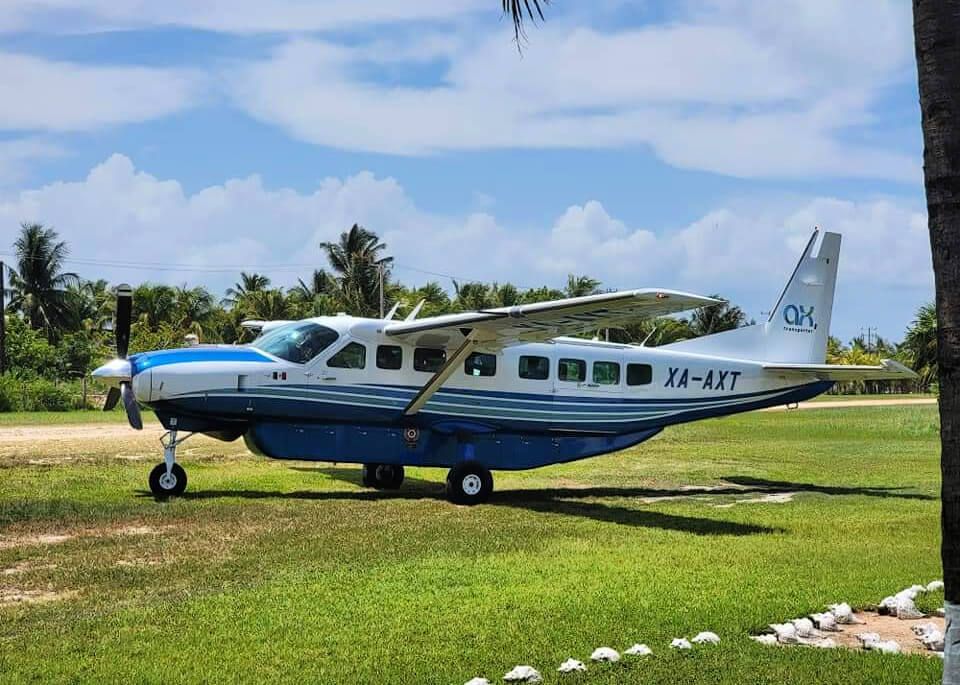 A blue and white small passenger airplane, with the registration "XA-AXT," parked on Holbox airstrip with lush palm trees in the background, having just completed a flight from Cancun to Holbox Island.