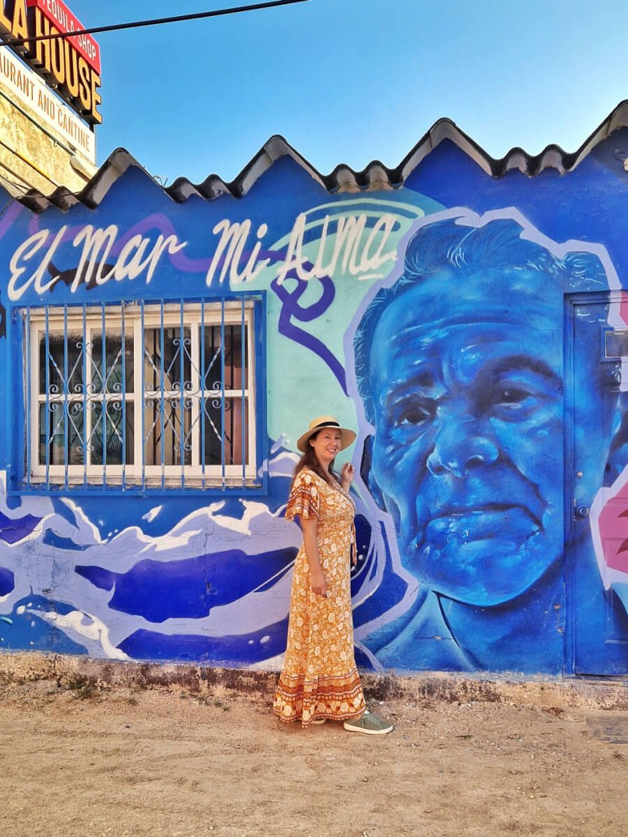 Sara in a patterned orange dress and a hat stands smiling to her side in front of a blue mural featuring a man's face and the text "El Mar Mi Alma" in Holbox, Mexico.