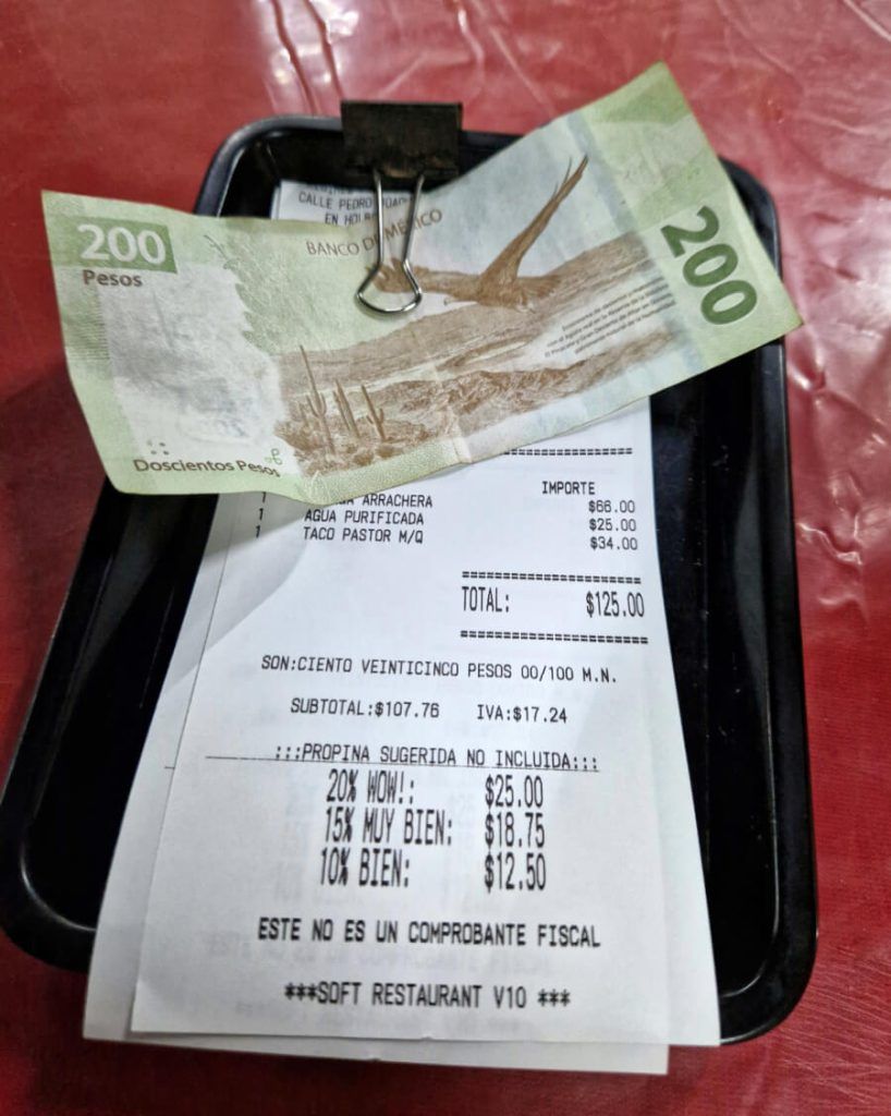 A 200 Mexican peso note clipped to a black bill holder, atop a receipt suggesting tip amounts of 20%, 15%, and 10% for services at a restaurant in Holbox.