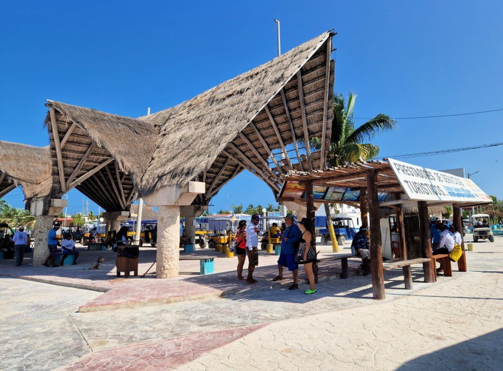 Thatched roof taxi stand in Holbox ferry port with parked golf cart taxis and tourists, under a clear blue sky.