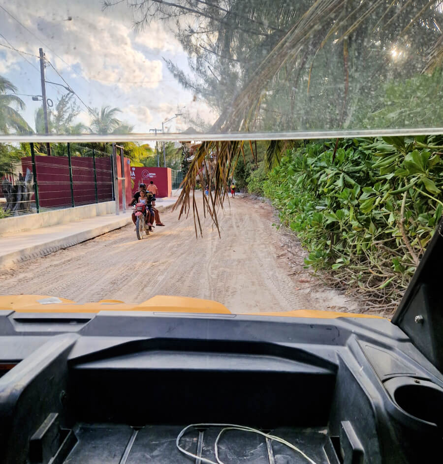 View from inside a golf cart taxi showing a motorcycle on a sandy road with tropical vegetation on the side in Holbox.
