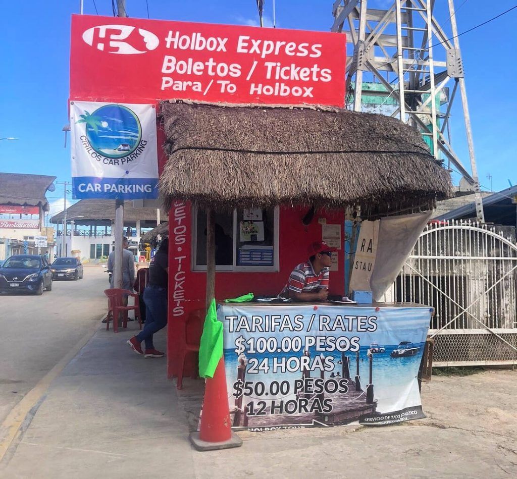 Ticket booth for Holbox Express ferry service and Chiquila car parking with posted rates, featuring a thatched roof, in a busy street setting.