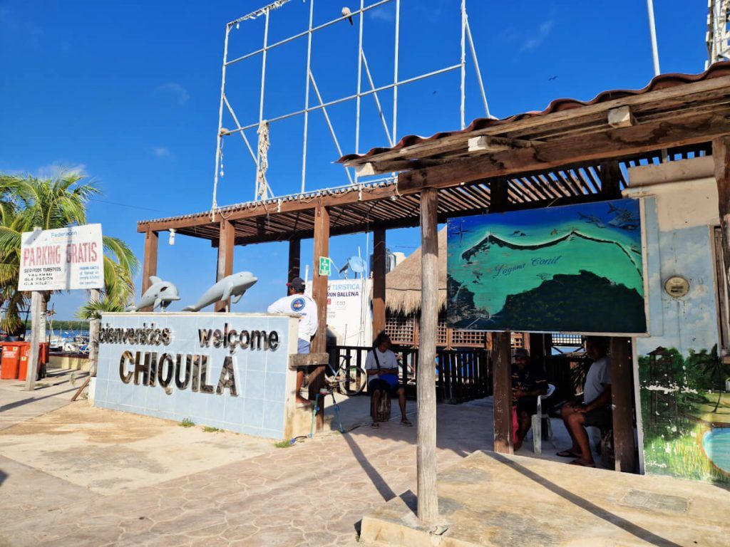 Welcoming entrance to Chiquila with a 'Bienvenidos Welcome CHIQUILA' sign, free parking sign, and people sitting under a shaded structure next to a map of Yucatan Peninsula.