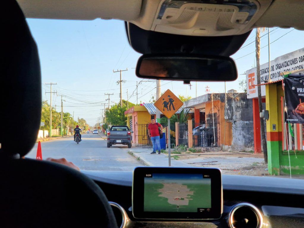 Interior perspective of driving through a town street from Cancun to Chiquila, with a school crossing sign ahead and a GPS on the dashboard.